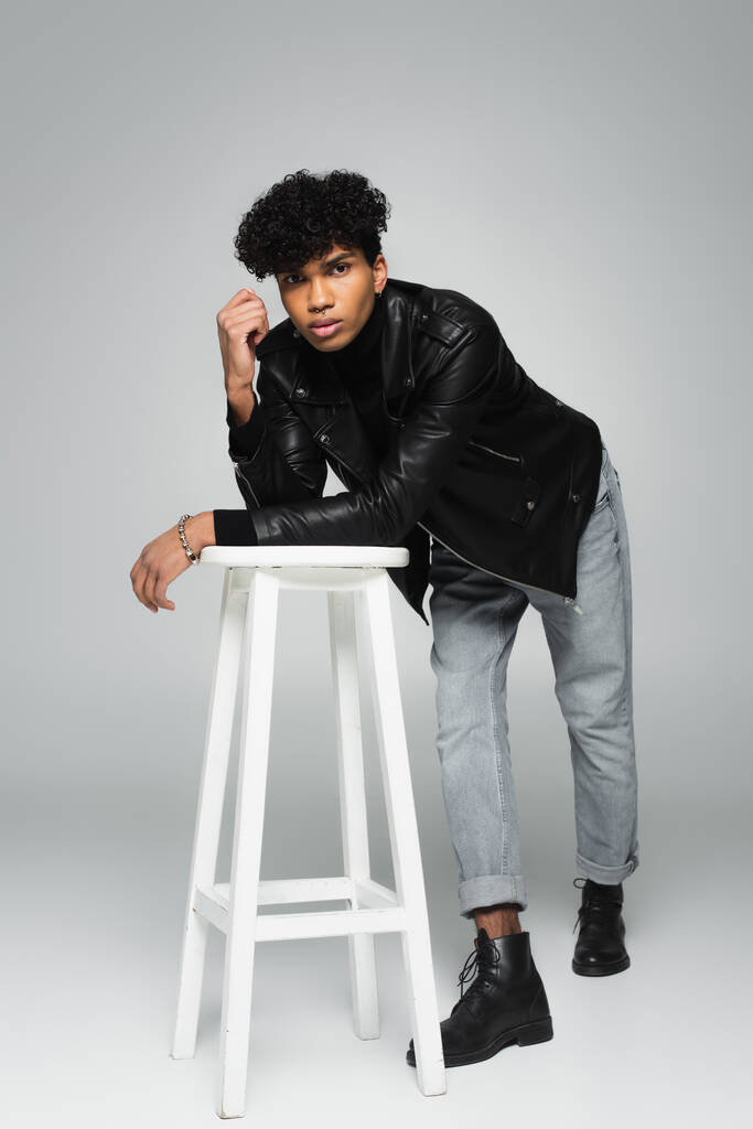 mladý africký Američan muž v černé kožené bundě a džíny opírající se o vysoké stoličce na šedém pozadí - Fotografie, Obrázek
