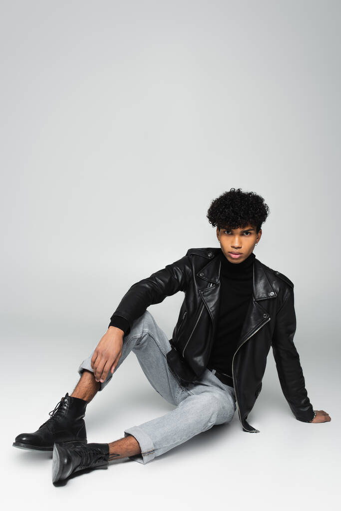 young african american man in black leather jacket and jeans sitting on grey background - Foto, Imagem