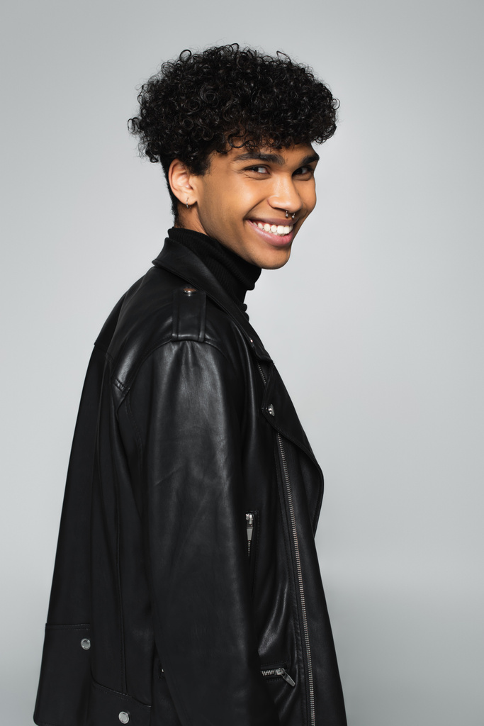 happy african american man in black leather jacket smiling at camera isolated on grey - 写真・画像