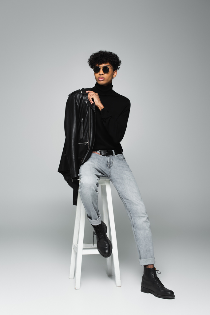 молодой африканский американец в стильной одежде и черных солнцезащитных очках, сидящий на высоком стуле на сером фоне - Фото, изображение