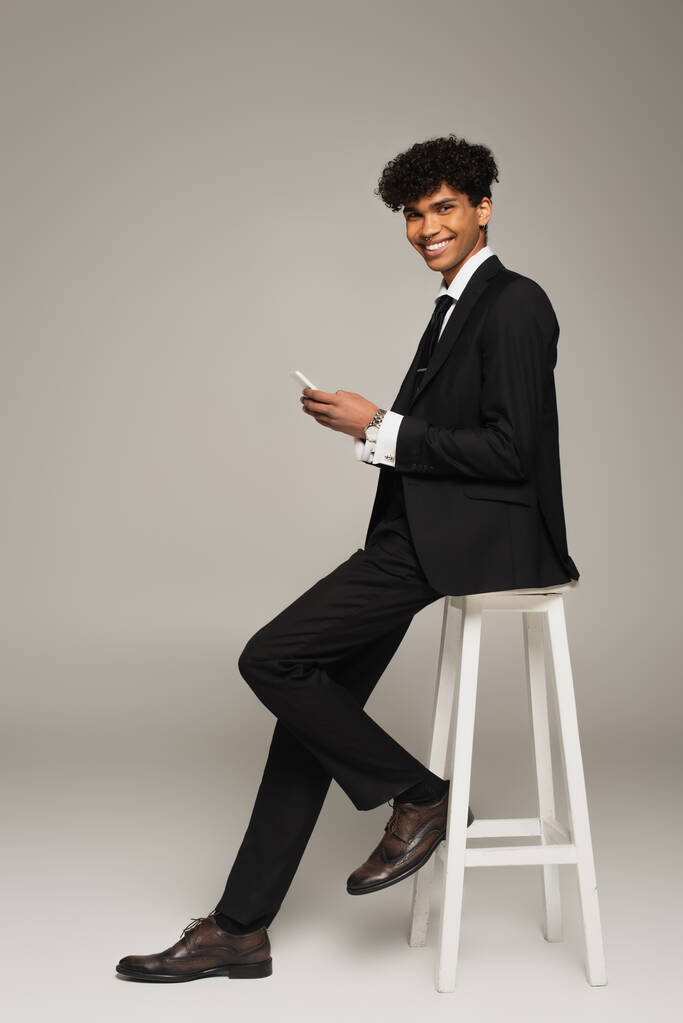 elegantní africký Američan s mobilním telefonem s úsměvem na kameru, zatímco sedí na šedém pozadí - Fotografie, Obrázek