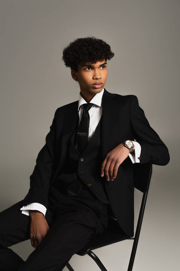 Афроамериканець у чорному костюмі з трьох частин сидить на стільці і озирається геть ізольовано на сірий - Фото, зображення