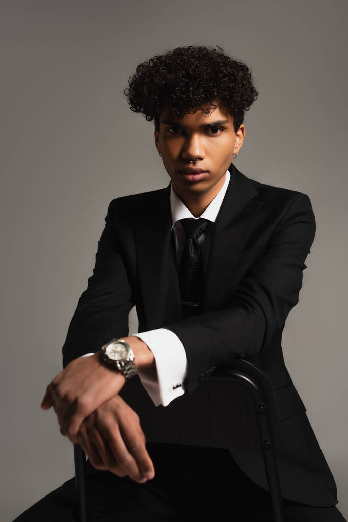 young african american businessman in black elegant suit sitting on chair and looking at camera isolated on grey - Photo, image