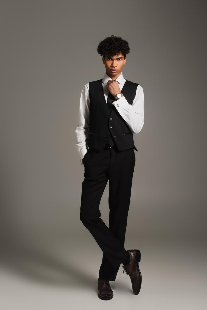 elegancki Afroamerykanin człowiek regulowanie czarny krawat podczas pozowanie z ręką w kieszeni na szarym tle - Zdjęcie, obraz