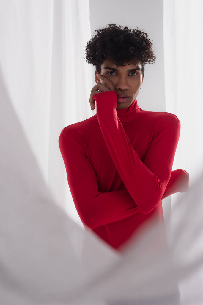 young african american man in red turtleneck posing with hand near face near white tulle cloth - Foto, Imagem