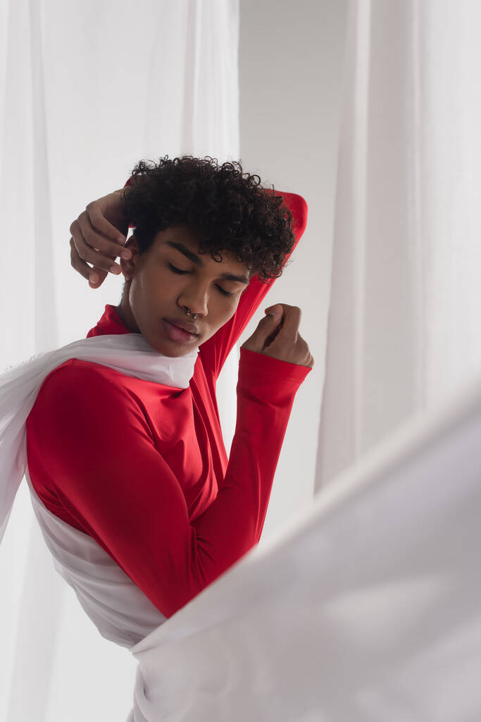 african american man in red turtleneck posing with closed eyes near white tulle drapery - 写真・画像