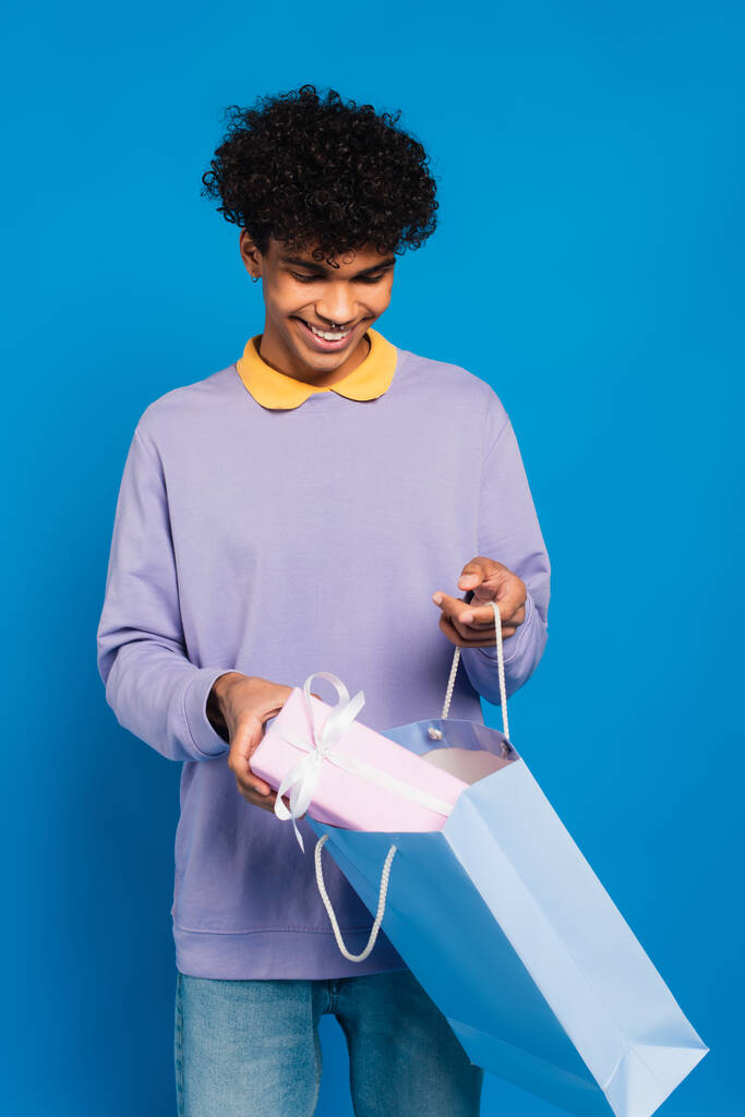cheerful african american man putting present into paper bag isolated on blue - Fotoğraf, Görsel
