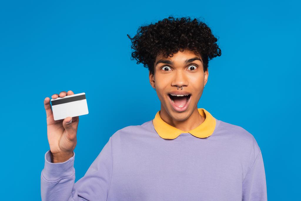 astonished african american man in violet pullover showing credit card isolated on blue - Foto, Imagem