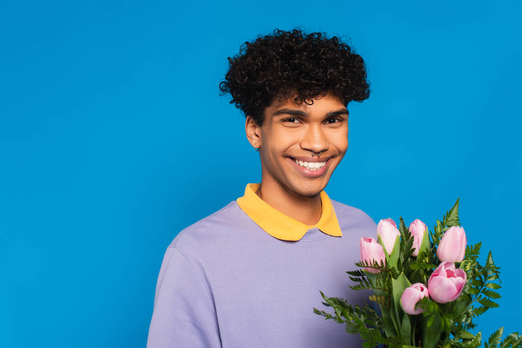 vidám afro-amerikai férfi rózsaszín tulipánnal mosolyog a kamera elszigetelt kék - Fotó, kép