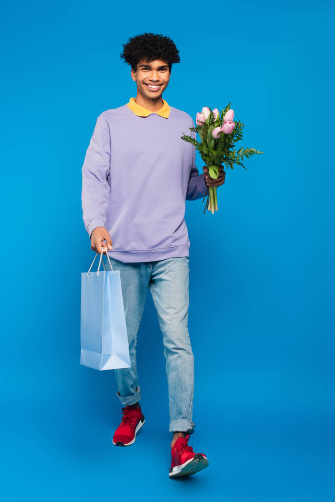 full length view of happy african american man walking with bouquet and shopping bag on blue background - Foto, Imagen