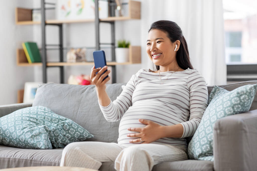 беременная женщина с телефоном и наушниками дома - Фото, изображение