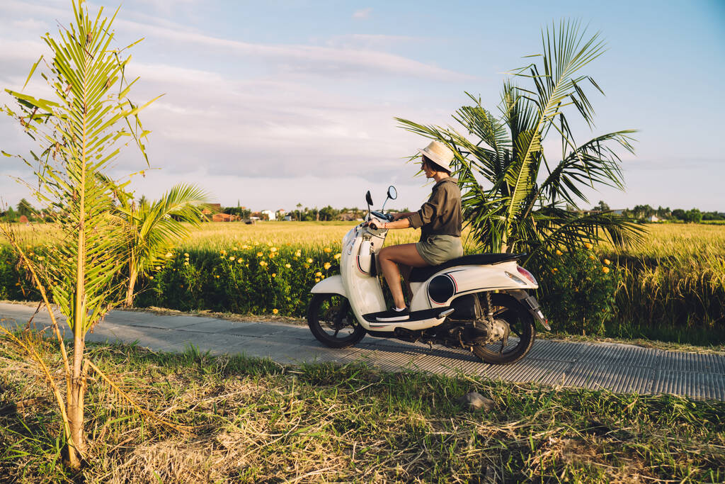 Vue latérale de la moto de conduite touristique féminine habillée décontractée explorant l'Indonésie pendant l'été pour le voyage d'escapade, jeune fille hipster arriver à destination sur Bali via scooter de location vintage - Photo, image