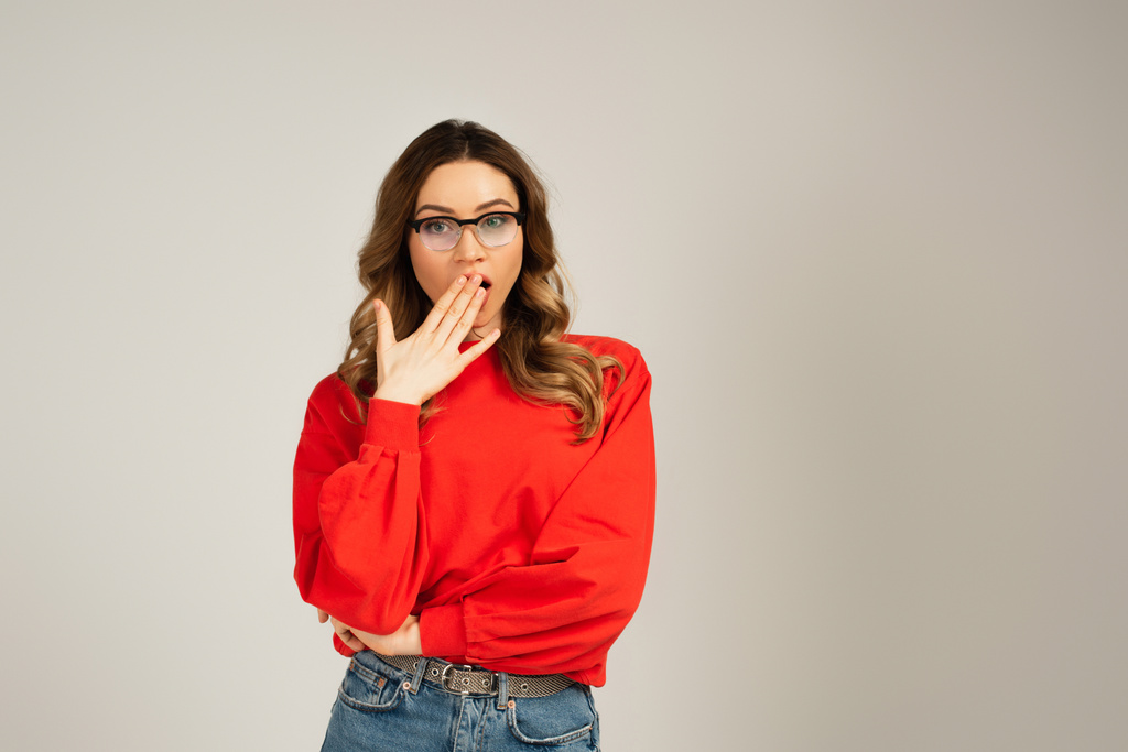 mulher chocada em camisola e óculos cobrindo boca isolada em cinza - Foto, Imagem
