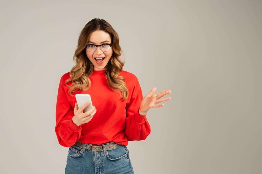 podekscytowana kobieta w okularach trzyma telefon komórkowy odizolowany na szaro - Zdjęcie, obraz