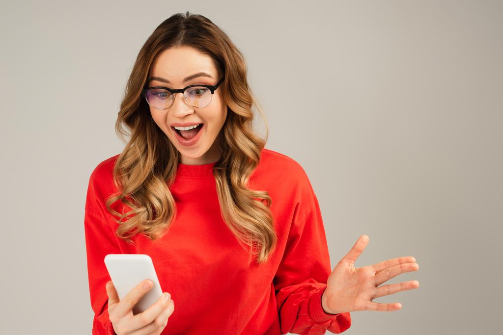 excited woman in eyeglasses looking at mobile phone isolated on grey - Photo, Image