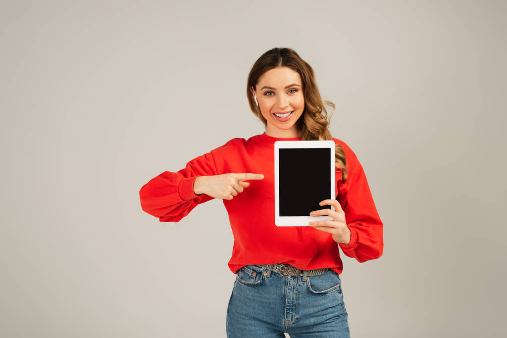 fröhliche Frau mit drahtlosen Kopfhörern, die auf ein digitales Tablet mit leerem Bildschirm zeigt, isoliert auf grau  - Foto, Bild