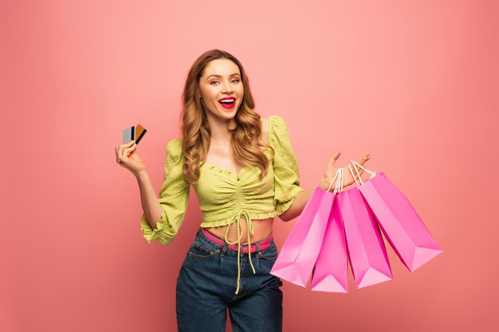 mujer sorprendida sosteniendo tarjetas de crédito y bolsas de compras aisladas en rosa  - Foto, imagen
