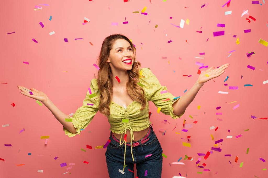 cheerful woman with outstretched hands near confetti on pink - Photo, Image
