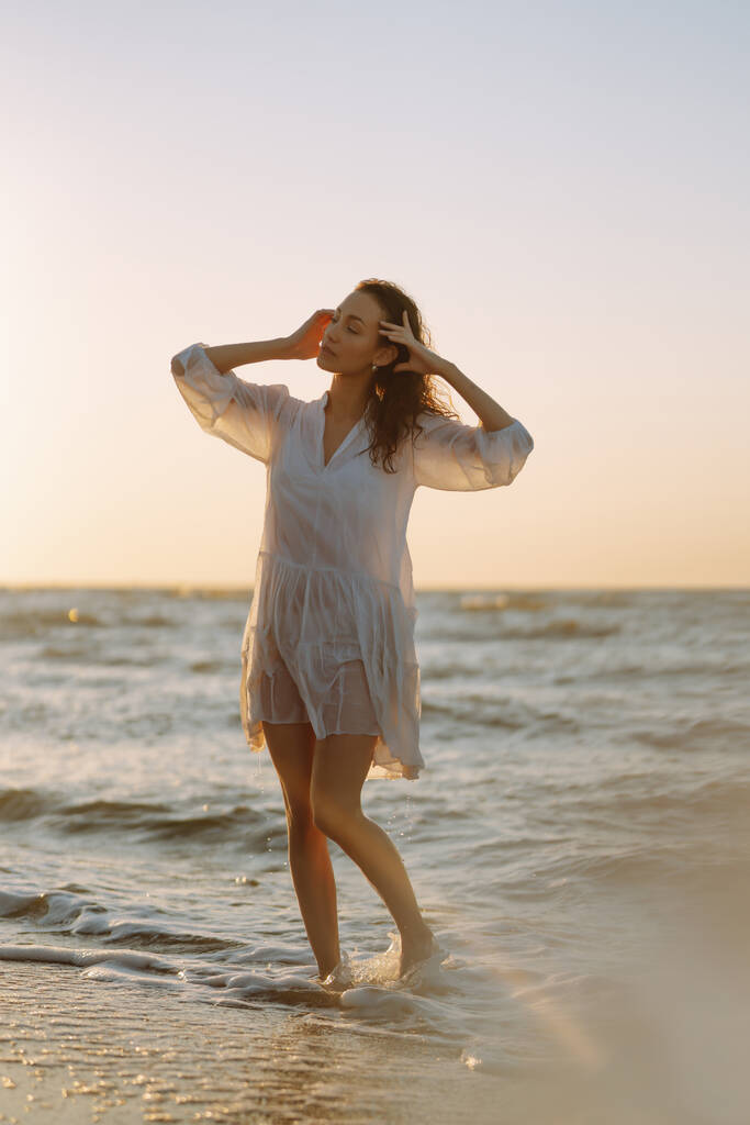Молодая счастливая женщина в белом трепещущем платье идет вдоль побережья. Девушка смотрит на волшебный восход солнца. Летнее время. Путешествия, выходные, отдых и образ жизни. - Фото, изображение