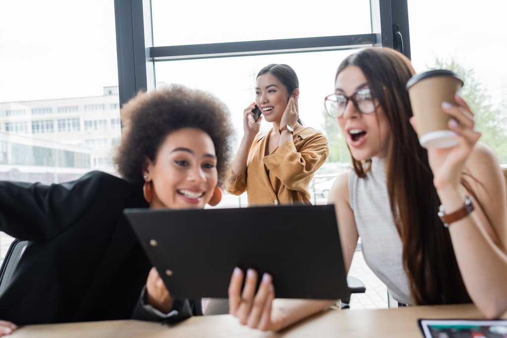 emocionado interracial managers mirando portapapeles cerca mientras feliz asiático mujer de negocios hablando en smartphone - Foto, Imagen