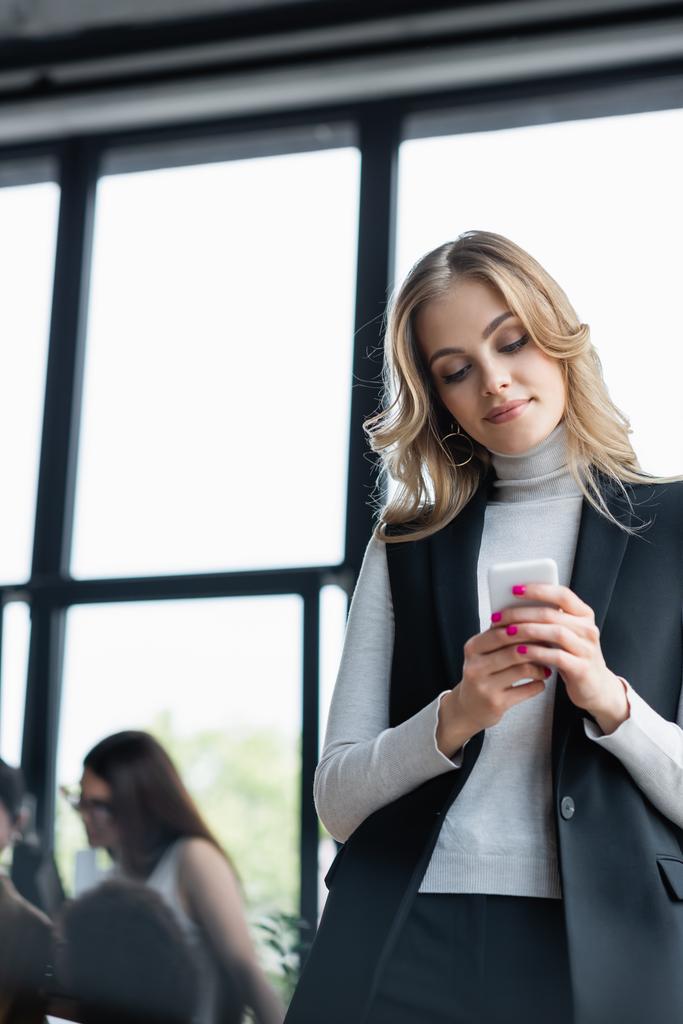 blonde femme d'affaires messagerie sur smartphone près de collègues sur fond flou - Photo, image