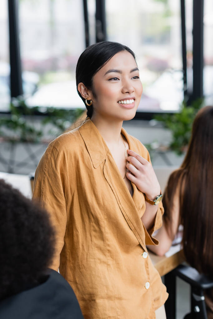 alegre asiático mujer de negocios mirando lejos mientras de pie cerca colegas en borrosa primer plano - Foto, imagen