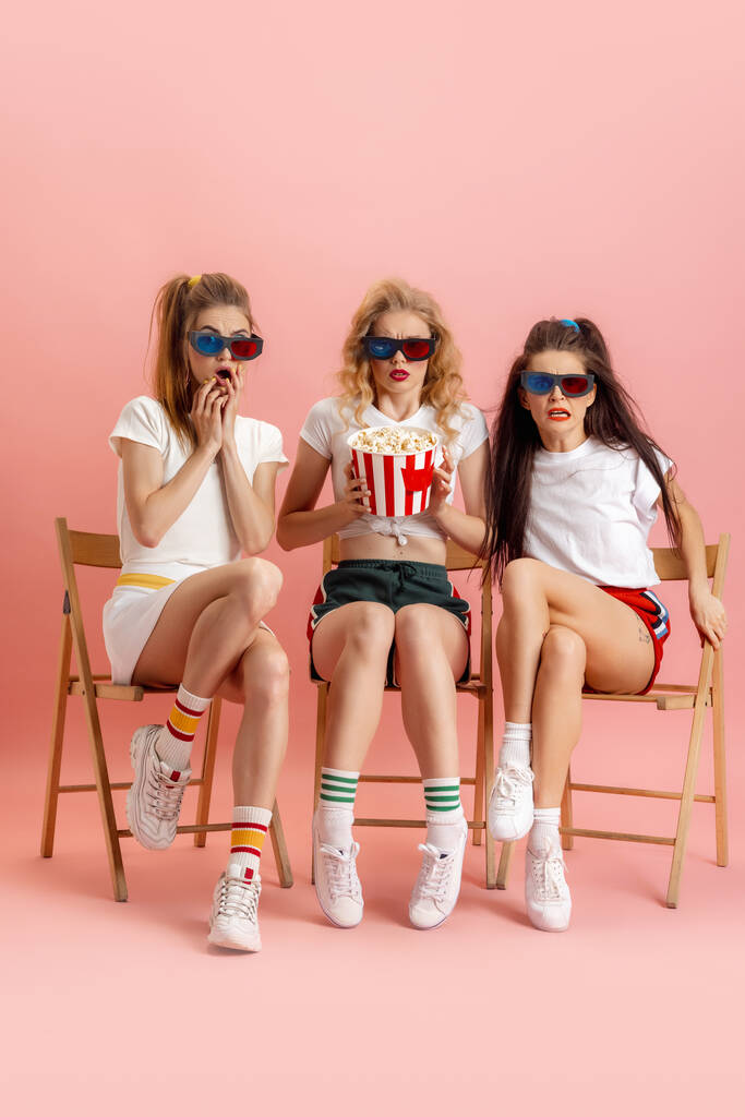 Τρεις νέες γυναίκες σε ρετρό στυλ μόδας της δεκαετίας του '90, ρούχα βλέποντας ταινία που απομονώνονται σε ροζ φόντο στούντιο. Έννοια της σύγκρισης εποχών, ομορφιάς, μόδας και νεολαίας. - Φωτογραφία, εικόνα
