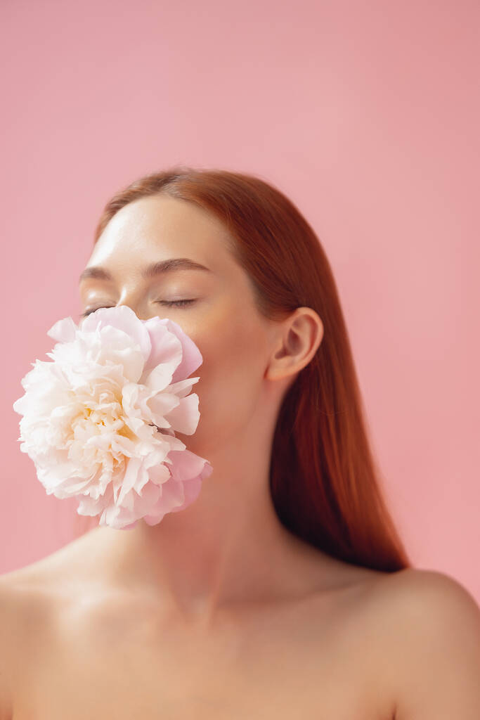 Detailní portrét krásné zrzavé ženy s květinou izolované přes růžové studio zázemí. Nahá barva, strava, kosmetika, přírodní krása a estetická kosmetologie. - Fotografie, Obrázek