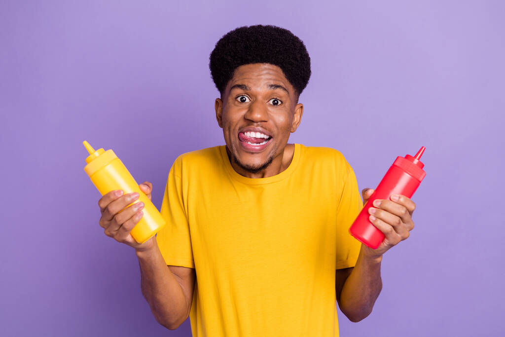 Retrato de chico alegre funky atractivo sosteniendo botellas de ketchup lamiendo bocadillo labial aislado sobre violeta color púrpura fondo - Foto, Imagen