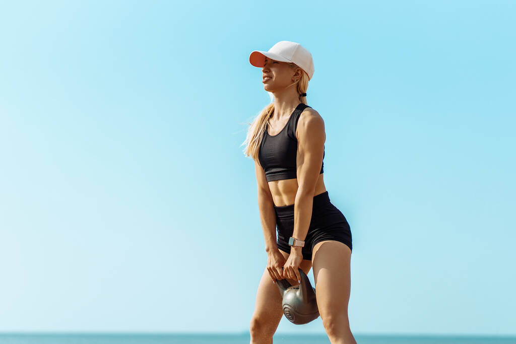 Spor giyimli spor elbiseli kadın ağırlık kaldırıyor, sportif kadın sabahları sahilde antrenman yaparken elinde kettlebell tutuyor. - Fotoğraf, Görsel
