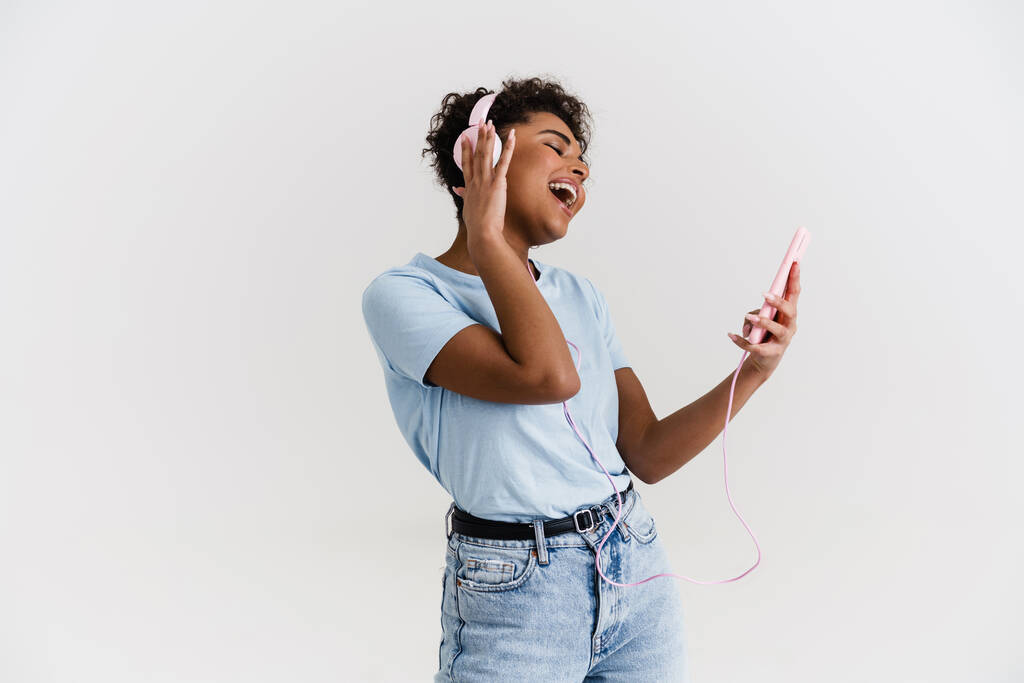 Смеющаяся черная женщина слушает музыку с наушниками и мобильным телефоном изолированы на белом фоне - Фото, изображение