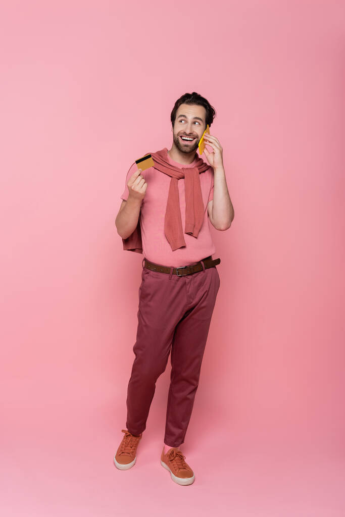 Улыбающийся мужчина разговаривает на смартфоне и держит кредитку во время покупок в интернете на розовом фоне  - Фото, изображение