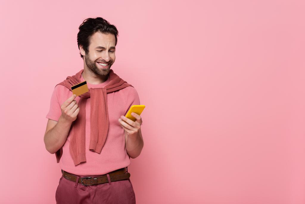 Позитивный человек держит кредитную карту и с помощью мобильного телефона изолированы на розовый  - Фото, изображение