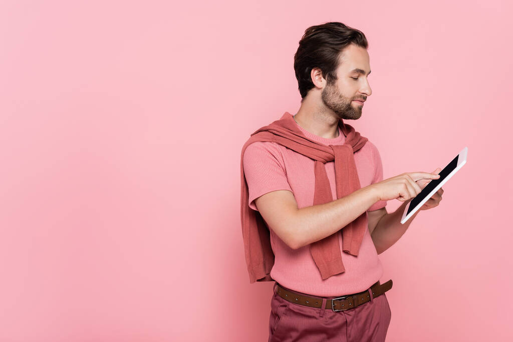 Вид сбоку на человека с джинсами на плечах с помощью цифровой таблички, изолированной на розовом фоне  - Фото, изображение