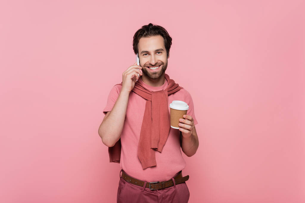Позитивный человек смотрит в камеру, разговаривая по мобильному телефону и держа кофе, чтобы пойти изолированы на розовый  - Фото, изображение