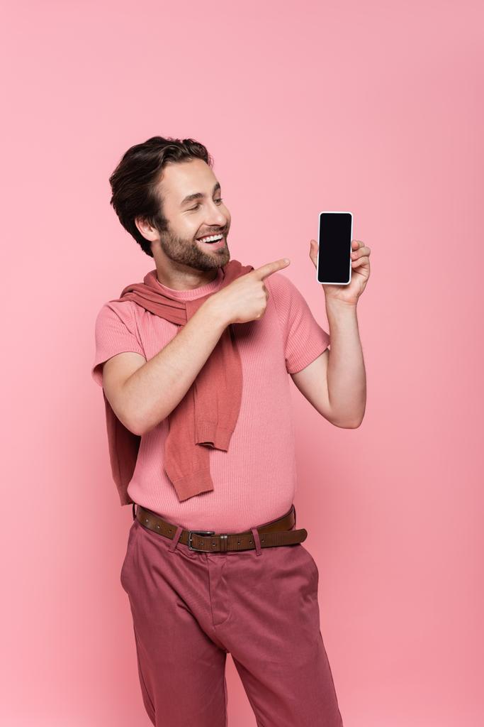 Usmívající se muž ukazuje na smartphone s prázdnou obrazovkou izolované na růžové  - Fotografie, Obrázek