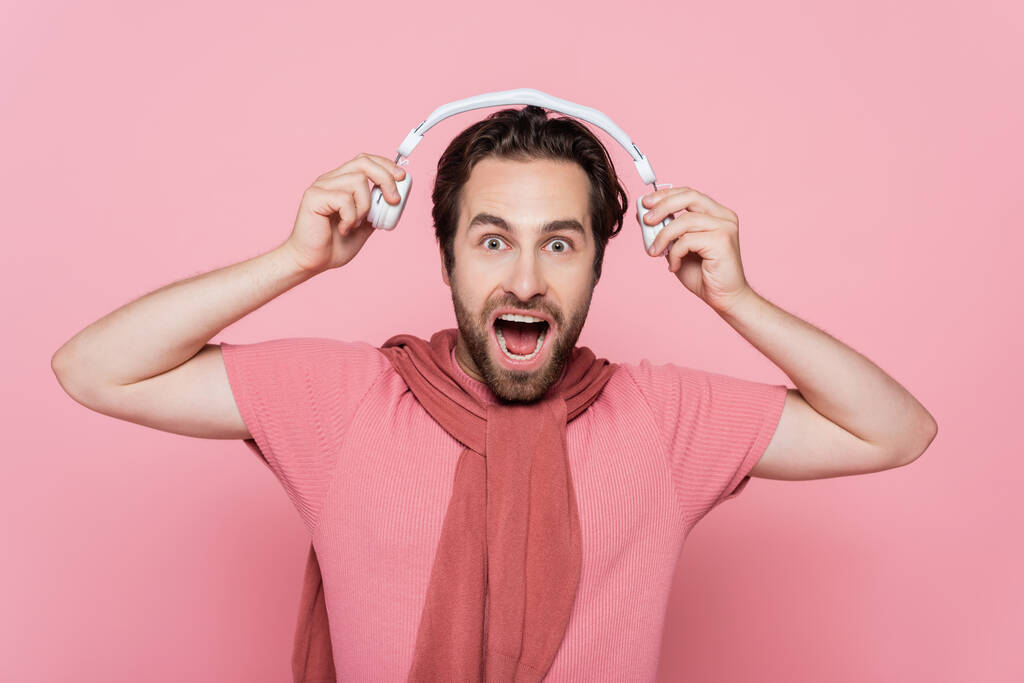 Amazed man with jumper on shoulders holding headphones isolated on pink  - Photo, Image