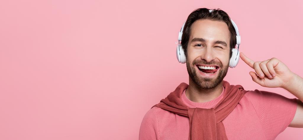 Χαμογελώντας άνθρωπος με ακουστικά να κλείνει το μάτι και να προεξέχει γλώσσα απομονωμένη σε ροζ, banner - Φωτογραφία, εικόνα