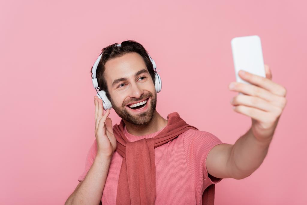 Smiling man in headphones taking selfie on blurred smartphone isolated on pink  - Photo, Image
