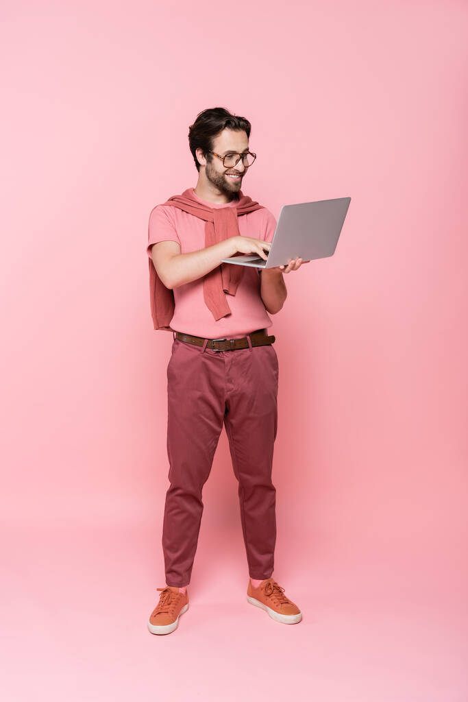 Longitud completa de freelancer sonriente usando portátil sobre fondo rosa - Foto, imagen