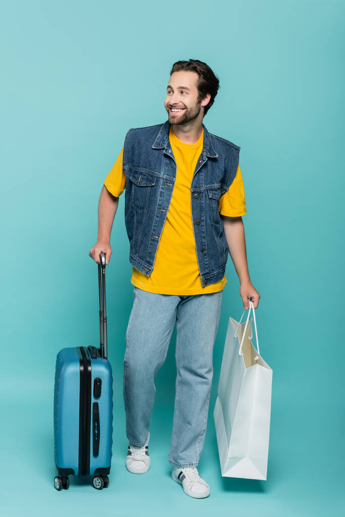Χαμογελαστός τουρίστας με βαλίτσα και τσάντα για ψώνια σε μπλε φόντο  - Φωτογραφία, εικόνα