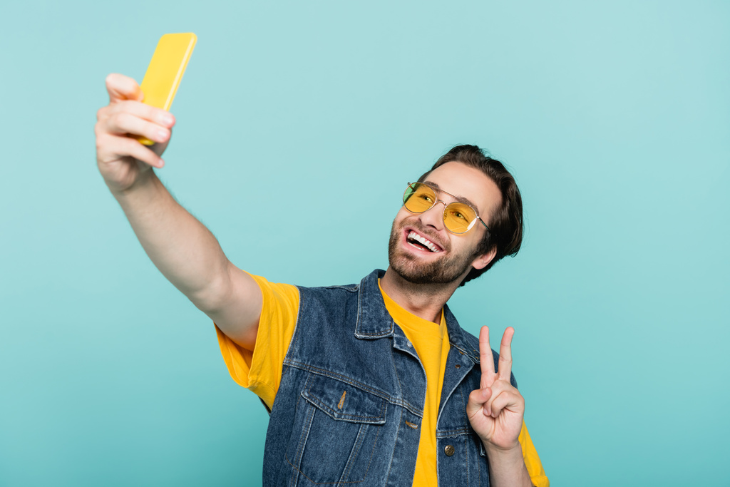Χαμογελώντας άνθρωπος με γιλέκο denim δείχνει σημάδι ειρήνης, ενώ τη λήψη selfie απομονώνονται σε μπλε  - Φωτογραφία, εικόνα