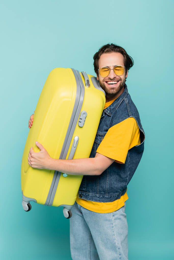 Homem feliz em óculos de sol abraçando mala isolada em azul  - Foto, Imagem
