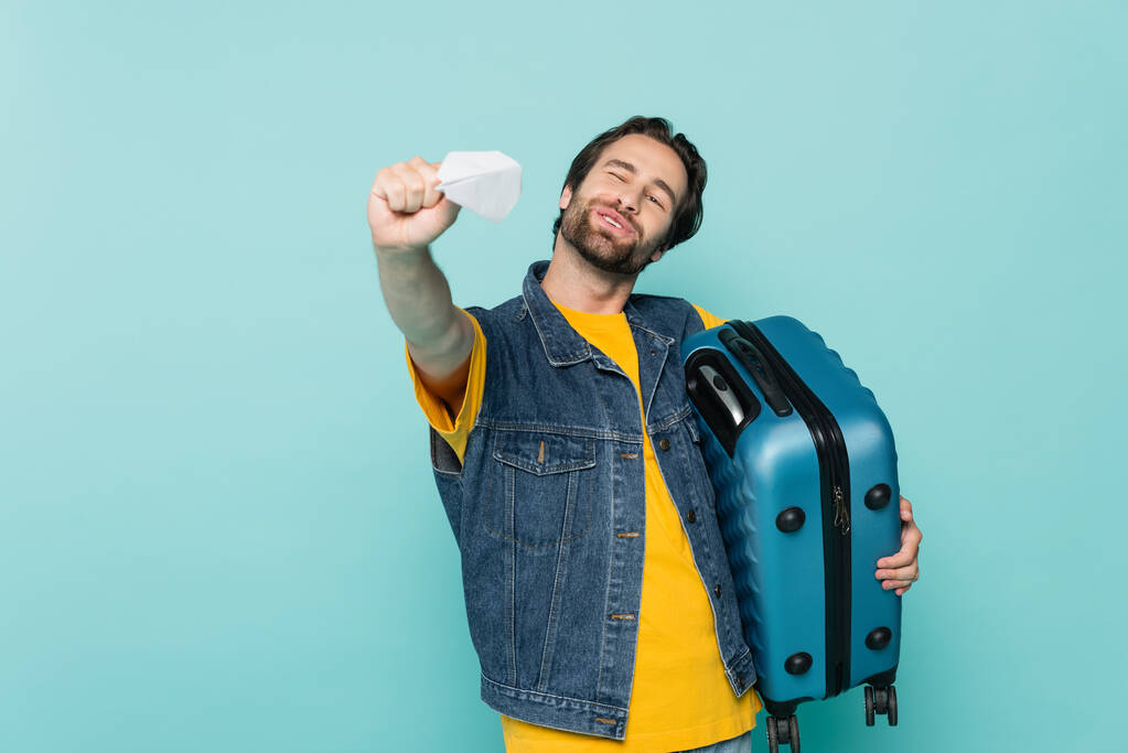 Positiivinen mies pitelee matkalaukkua ja leikkii sinisellä eristetyllä paperilentokoneella  - Valokuva, kuva