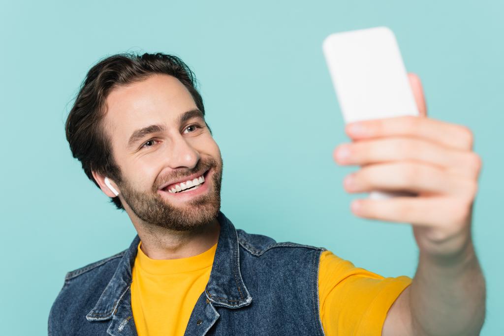 Homme souriant dans l'écouteur prendre selfie sur smartphone isolé sur bleu  - Photo, image