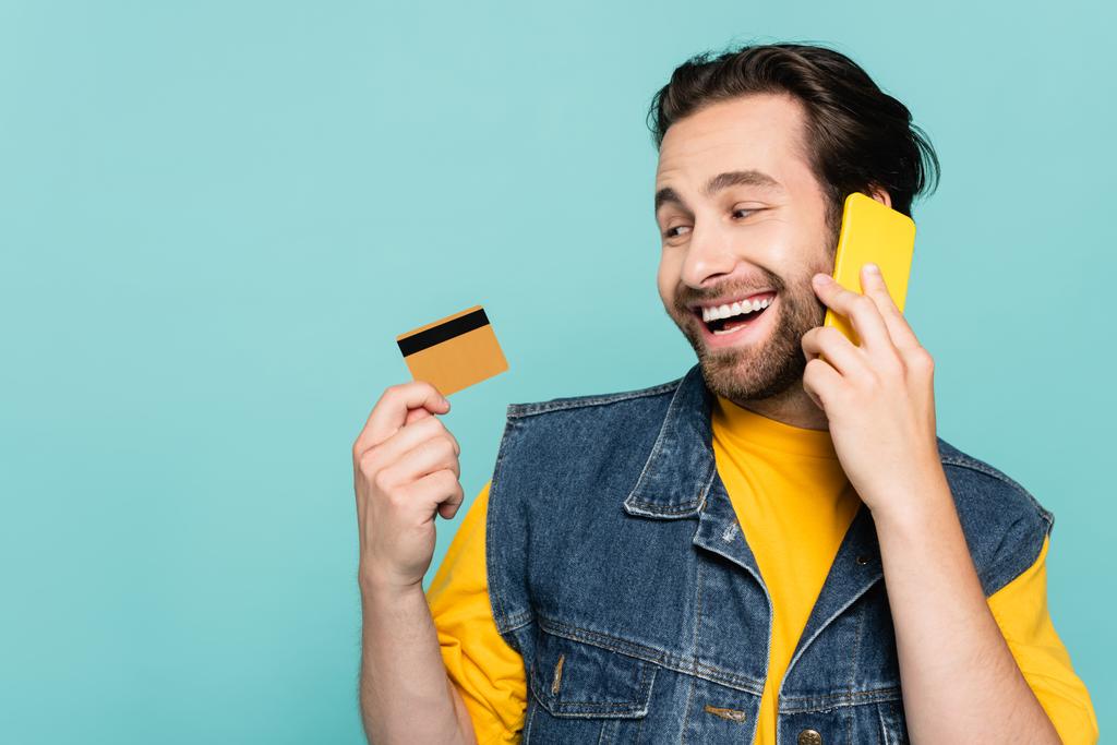 Uśmiechnięty mężczyzna rozmawiający przez telefon komórkowy i patrzący na kartę kredytową odizolowaną na niebiesko - Zdjęcie, obraz