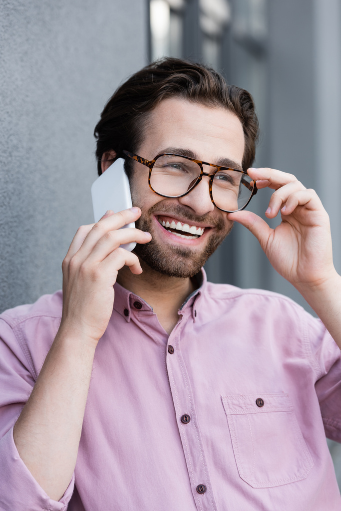 Улыбающийся бизнесмен, держащий очки, разговаривая по смартфону возле здания  - Фото, изображение
