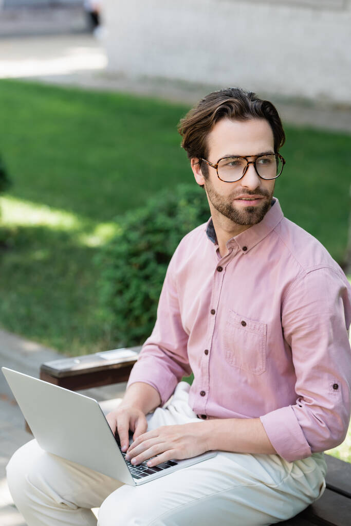 Бізнесмен в окулярах використовує ноутбук на лавці на відкритому повітрі
  - Фото, зображення