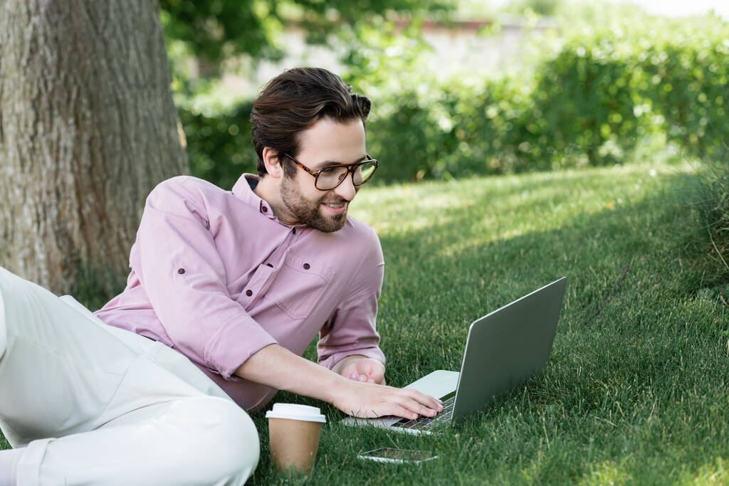 Businessman using laptop near coffee to go and smartphone on grass in park  - Photo, Image