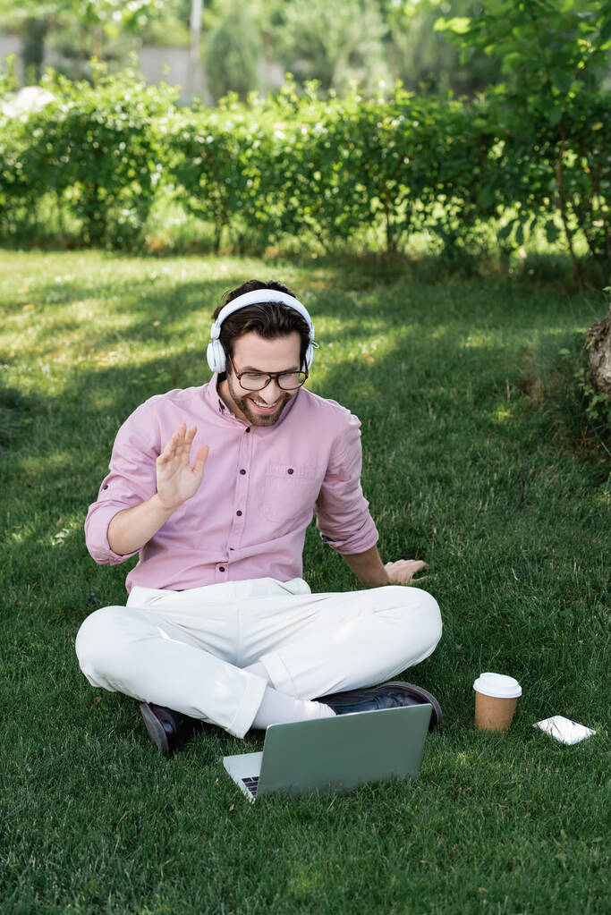 Homme d'affaires souriant dans les écouteurs ayant un appel vidéo sur ordinateur portable près de tasse de papier sur l'herbe dans le parc  - Photo, image